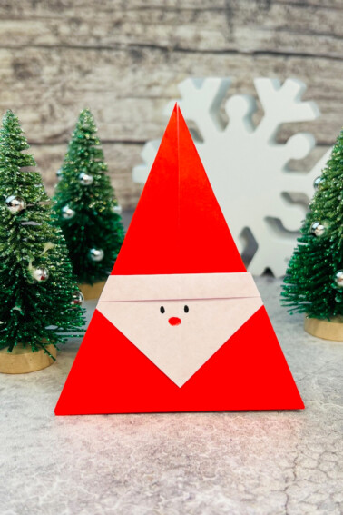 Image shows a finished origami Santa with small Christmas trees in the background. KidsActivitiesBlog